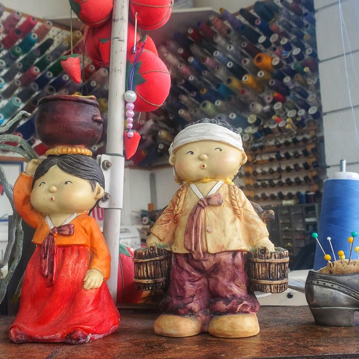 Two small ceramic figurines in front of a wall of colorful thread spools and pincushions at the alterations counter